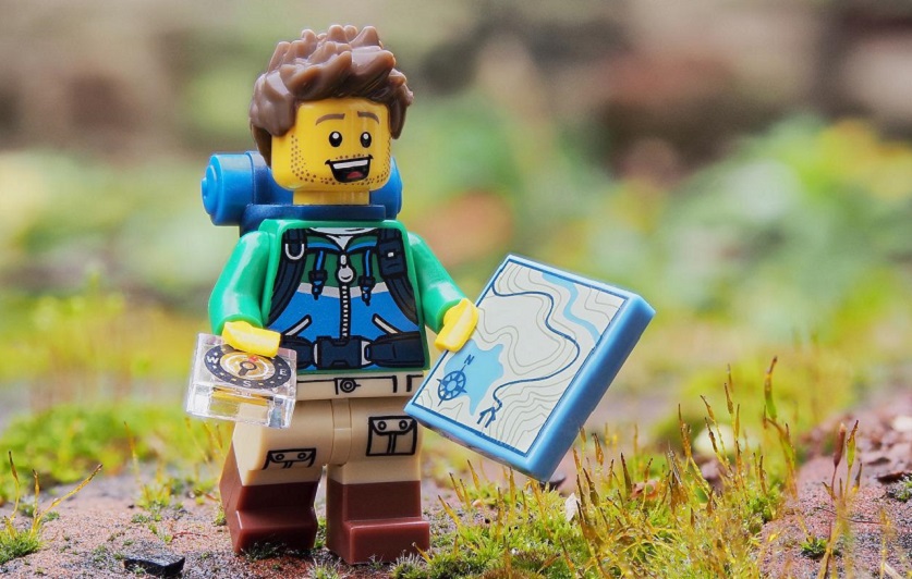 Un randonneur en légo portant une carte et une boussole. Il a une barbe de trois jours. Il est posé sur un rocher avec de l'herbe.